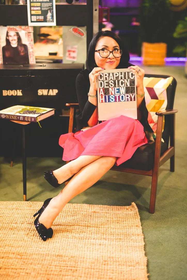 Rebecca Leppard seated in a pink skirt, holding the book Graphic Design: A New History