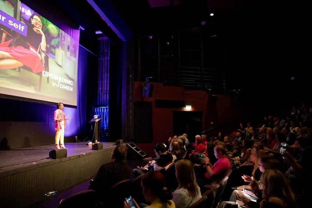Rebecca on a theatre stage beneath an Upgrading Women screen, facing a full auditorium